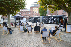 Auftaktveranstaltung "DemokratieWagen/StreitBus" aus dem Hintergrund fotografiert. Zu sehen sind Menschen, die in Liegestühlen sitzen und einer Rede zuhören. Die Rednerin steht vor dem Streitbus, der vor der Frankfurter Paulskirche steht.