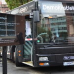 Frontalaufnahme des Demokratiewagens. Der Bus ist schwarz mit weißen Schrift- und Bildelementen. Links daneben steht ein Bildschirm, der das Programm des Veranstaltungstages anzeigt.