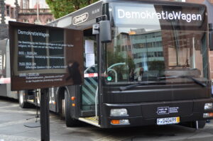 Frontalaufnahme des Demokratiewagens. Der Bus ist schwarz mit weißen Schrift- und Bildelementen. Links daneben steht ein Bildschirm, der das Programm des Veranstaltungstages anzeigt.